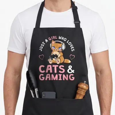 Cat gaming apron - Cartoon brown cat with headset holding controller between two pink hearts under arched text — shown on a black cotton canvas apron against a white background