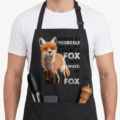 Alway fox aprons - Cartoon fox illustration next to stacked text reading ALWAYS BE YOURSELF UNLESS YOU CAN BE A FOX — shown on a black cotton canvas apron against a white background