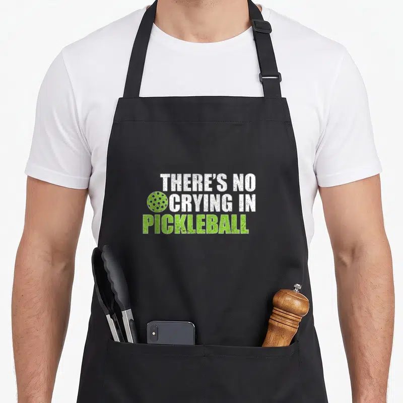 Crying pickleball apron - Stacked white and lime green distressed text with a pickleball graphic replacing a letter O — shown on a black cotton canvas apron against a white background