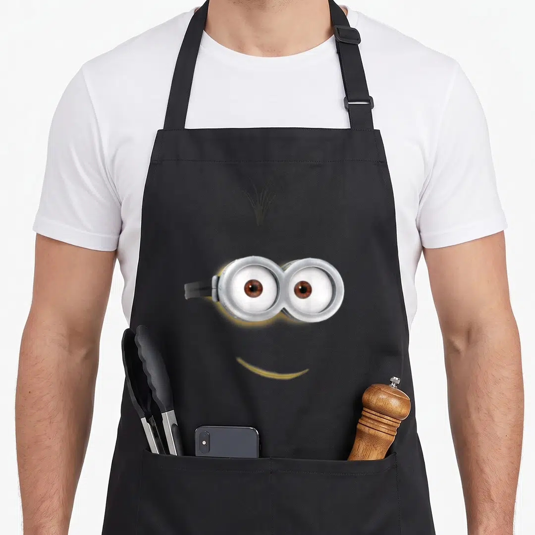 Despicable face on apron - Minimalist minion face with silver goggles, black hair tuft, and yellow smile curve — shown on a black cotton canvas apron against a white background