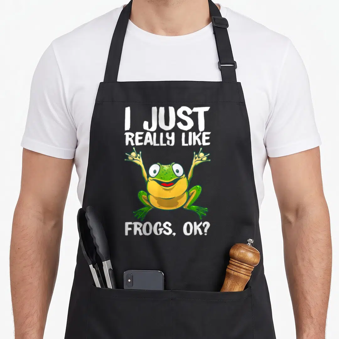 Tadpole frog aprons - Cartoon frog with raised arms between text I Just Really Like Frogs Ok in white — shown on a black cotton canvas apron against a white background