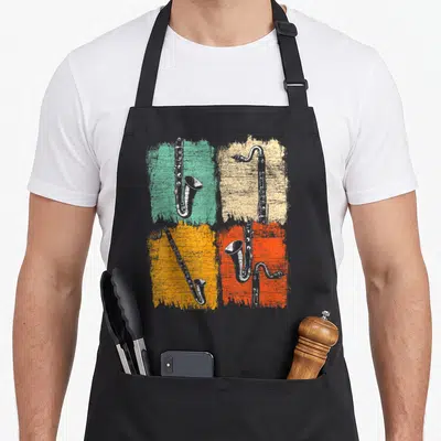 Bass clarinet life apron - Four white bass clarinets on distressed teal, beige, orange, and red-orange squares — shown on a black cotton canvas apron against a white background