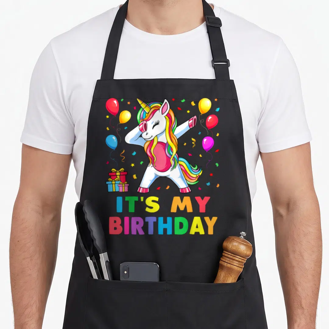 Cute unicorn birthday apron - Dabbing unicorn with balloons and IT'S MY BIRTHDAY text in rainbow colors — shown on a black cotton canvas apron against a white background
