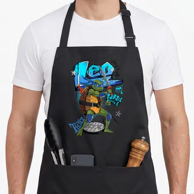 Mayhem teenage mutant ninja apron - Leonardo standing on manhole cover with LEO and TURTLE POWER text and scattered — shown on a black cotton canvas apron against a white background