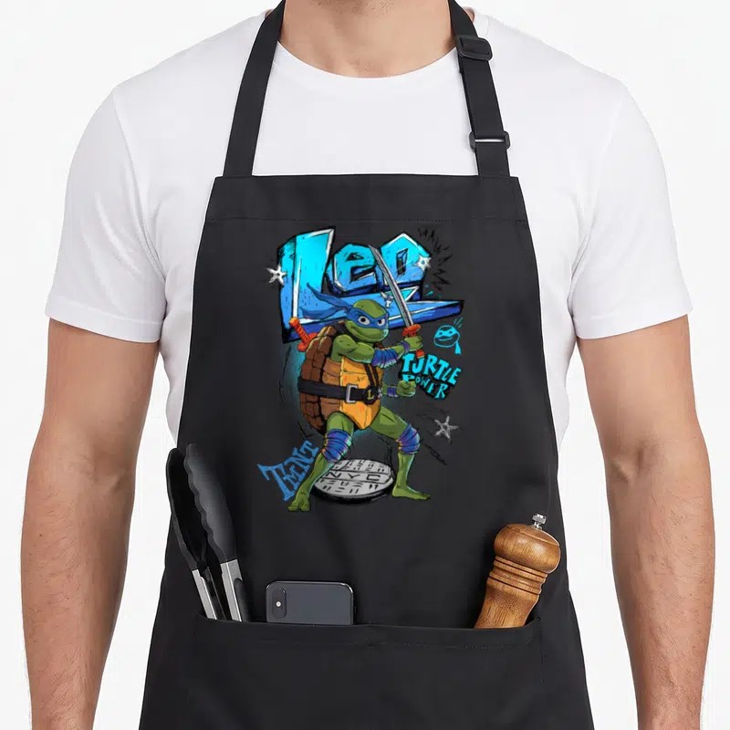 Mayhem teenage mutant ninja apron - Leonardo standing on manhole cover with LEO and TURTLE POWER text and scattered — shown on a black cotton canvas apron against a white background