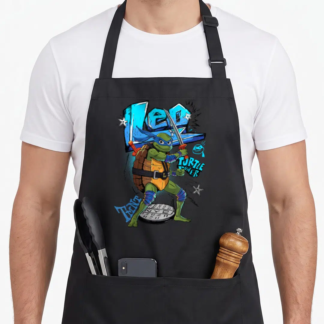 Mayhem teenage mutant ninja apron - Leonardo standing on manhole cover with LEO and TURTLE POWER text and scattered — shown on a black cotton canvas apron against a white background