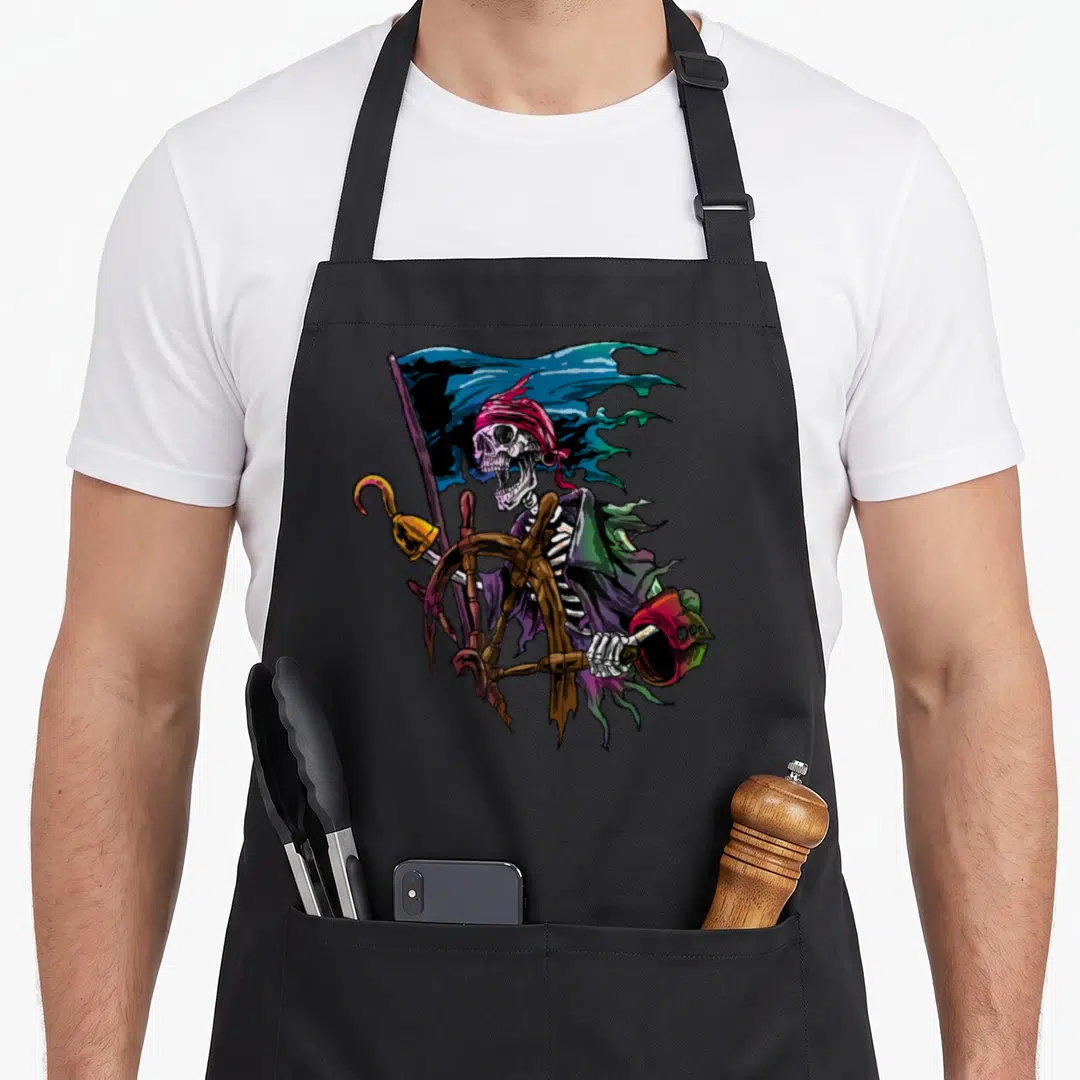 Sea skull apron - Cartoon skeleton pirate holding ship wheel and hook with tattered flag in purple green blue red — shown on a black cotton canvas apron against a white background