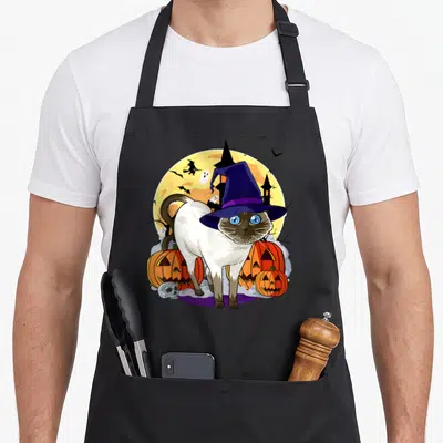 Halloween cute cat aprons - Siamese cat in purple witch hat with pumpkins, skull, and yellow moon with flying witch — shown on a black cotton canvas apron against a white background