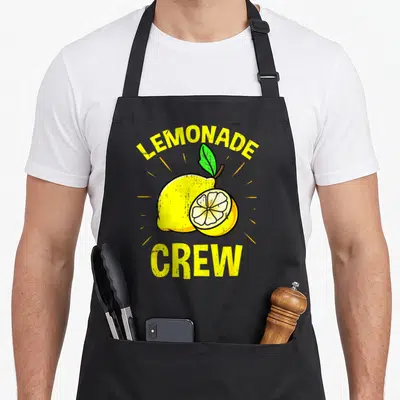 Funny crew lemonade apron - Arched LEMONADE CREW text over yellow cartoon lemon and slice with green leaf — shown on a black cotton canvas apron against a white background