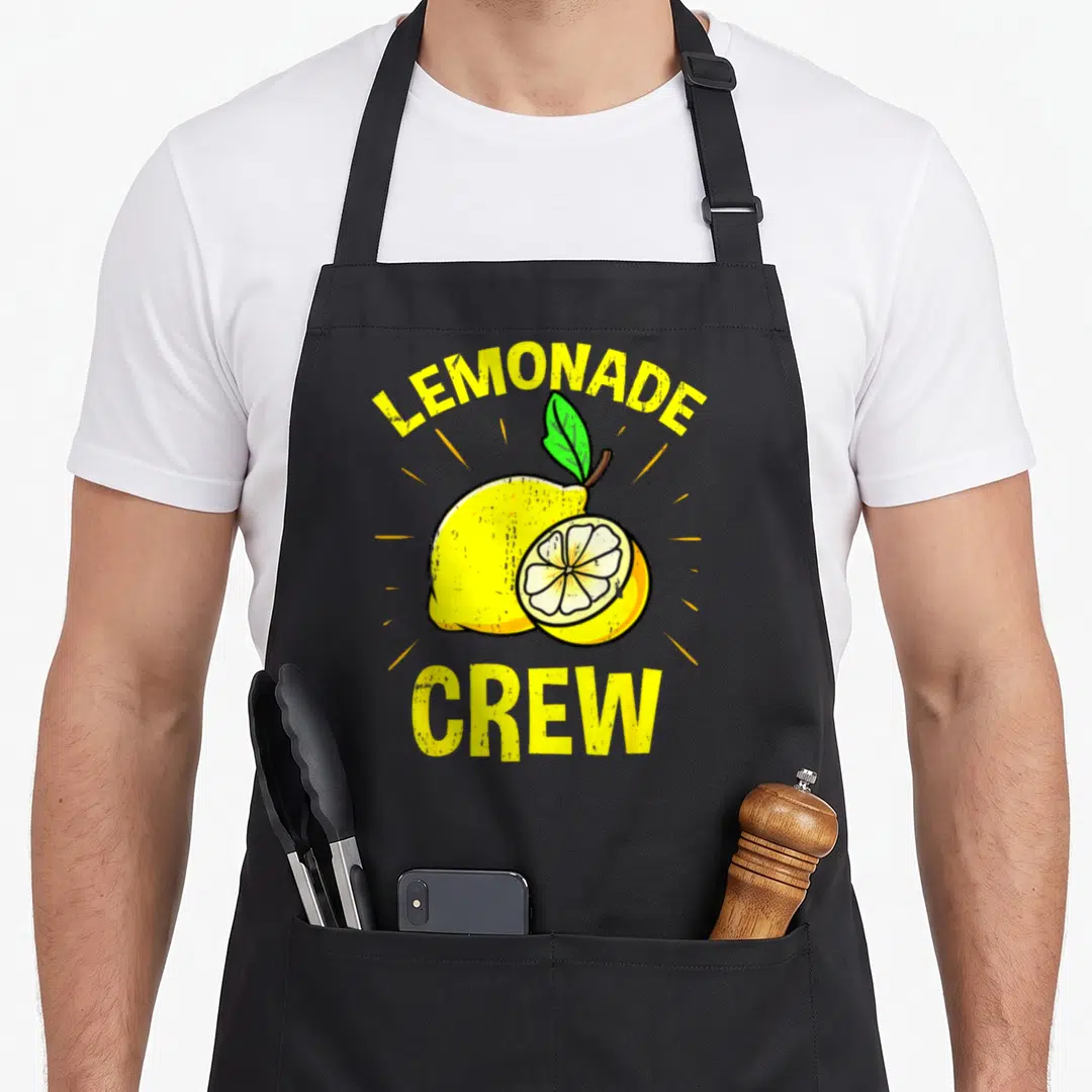 Funny crew lemonade apron - Arched LEMONADE CREW text over yellow cartoon lemon and slice with green leaf — shown on a black cotton canvas apron against a white background