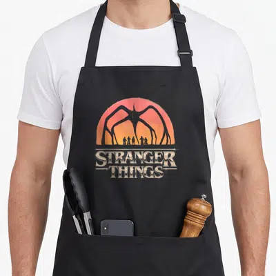 Season distressed apron - Grey monster silhouette over red orange sunset with Stranger Things text and bike riders — shown on a black cotton canvas apron against a white background