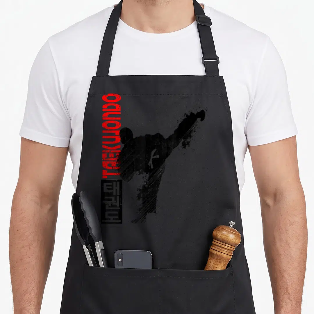 Martial distressed apron - Vertical TAEKWONDO text and black kicking figure silhouette with Korean characters — shown on a black cotton canvas apron against a white background