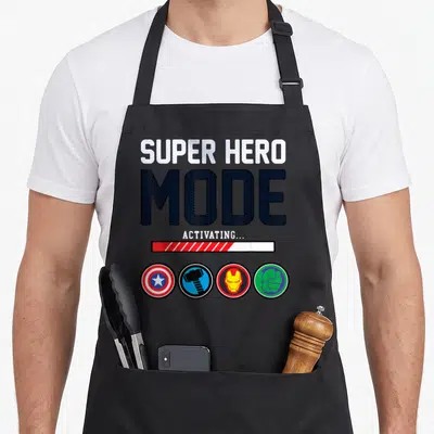 Mode super hero aprons - SUPER HERO MODE ACTIVATING text with loading bar and four circular icons in blue red and green — shown on a black cotton canvas apron against a white background