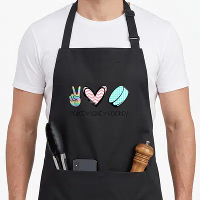 Peace wintersport hockey apron - Peace sign heart and puck with text in rainbow chevron and gingham patterns — shown on a black cotton canvas apron against a white background