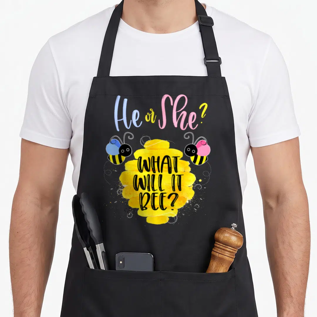 Gender reveal she bee apron - Watercolor bees in blue and pink flank a yellow honeycomb with 'What Will It Bee' text — shown on a black cotton canvas apron against a white background