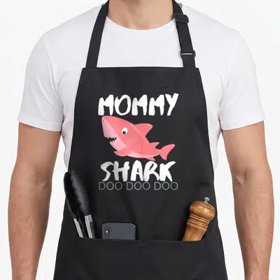 Shark mother's day aprons - Pink cartoon shark graphic with MOMMY SHARK DOO DOO DOO text in white and grey — shown on a black cotton canvas apron against a white background