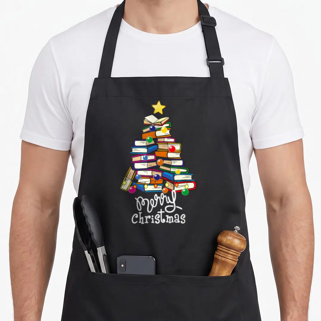 Merry christmas tree apron - Stacked books forming a tree shape with ornaments and white text below — shown on a black cotton canvas apron against a white background