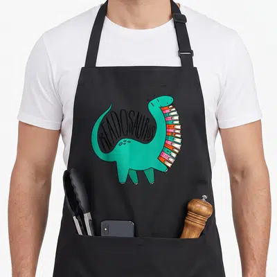 Readosaurus dinosaur apron - Teal cartoon dinosaur with book stack belly and curved READOSAURUS text — shown on a black cotton canvas apron against a white background