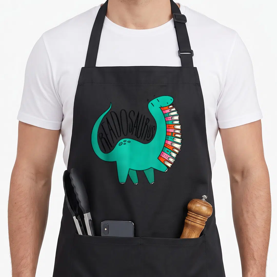 Readosaurus dinosaur apron - Teal cartoon dinosaur with book stack belly and curved READOSAURUS text — shown on a black cotton canvas apron against a white background