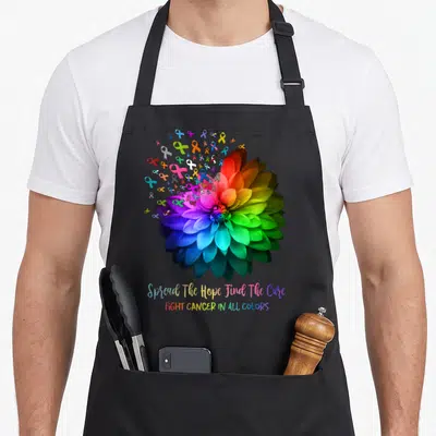 Fight find aprons - Realistic rainbow flower surrounded by red yellow green blue purple ribbons with hope cure text — shown on a black cotton canvas apron against a white background