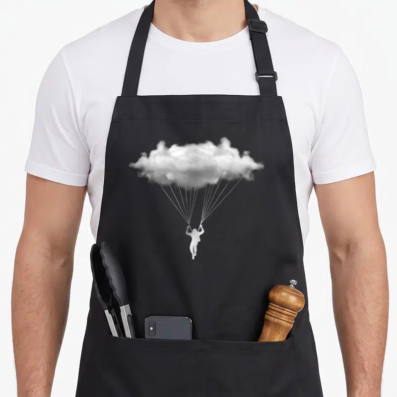 Cloud paragliding apron - White skydiver suspended by cords from a cloud acting as a parachute in photo style — shown on a black cotton canvas apron against a white background