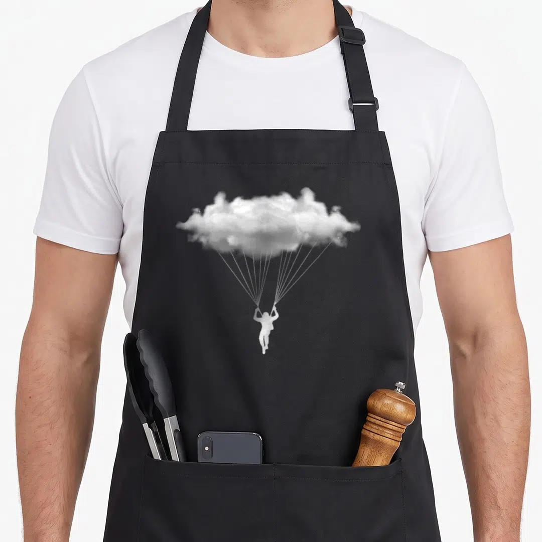 Cloud paragliding apron - White skydiver suspended by cords from a cloud acting as a parachute in photo style — shown on a black cotton canvas apron against a white background