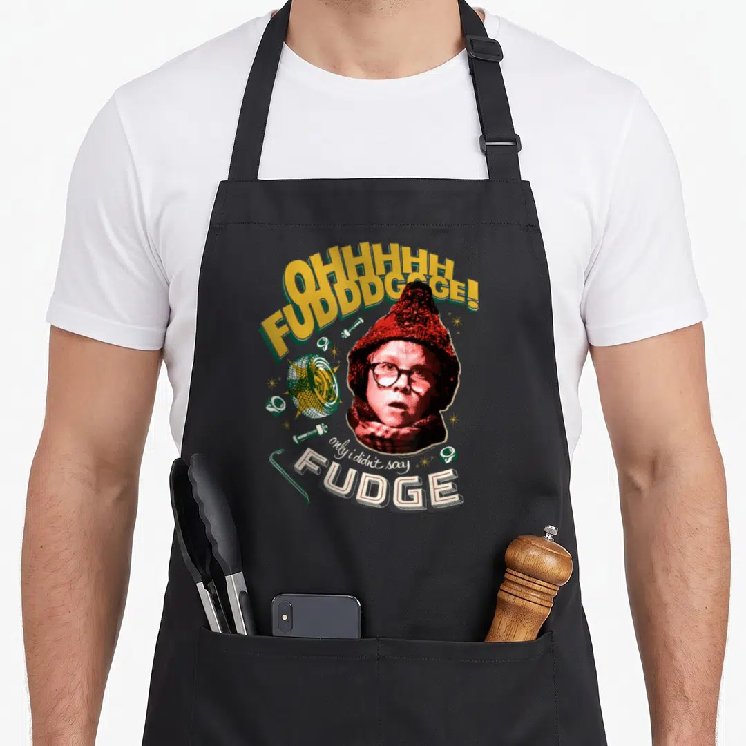 Story christmas apron - Retro boy face surrounded by yellow text OHHHHHHH FUDDDGGGE and scattered bolts in red green — shown on a black cotton canvas apron against a white background