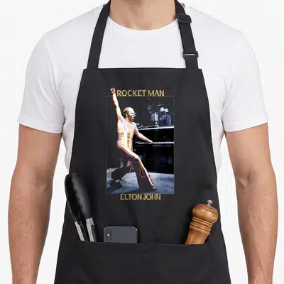 Elton x apron - Photo of man standing at piano in pink pants with gold stripes, framed by ROCKET MAN and ELTON JOHN text — shown on a black cotton canvas apron against a white background