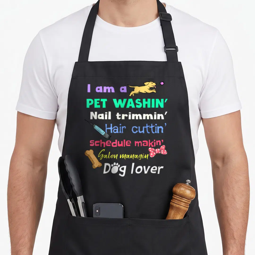 Groomer pet funny dog apron - Stacked colorful text listing grooming tasks with cartoon dog clipper and bone icons — shown on a black cotton canvas apron against a white background