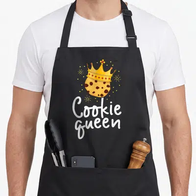 Chocolate cookie queen apron - Crowned cookie illustration with white Cookie Queen text and yellow sparkles — shown on a black cotton canvas apron against a white background