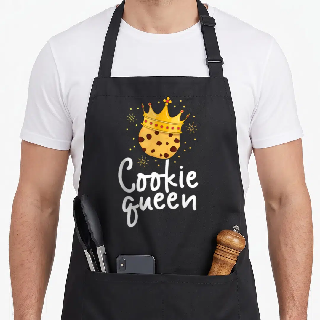 Chocolate cookie queen apron - Crowned cookie illustration with white Cookie Queen text and yellow sparkles — shown on a black cotton canvas apron against a white background