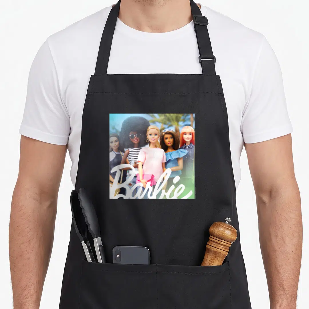 International tree apron - Five dolls standing side-by-side with white Barbie script overlay in photo style — shown on a black cotton canvas apron against a white background