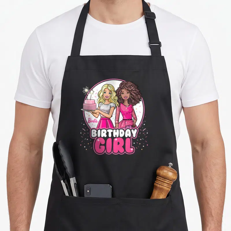 General bday apron - Two cartoon girls in pink circle holding cake with BIRTHDAY GIRL text and confetti — shown on a black cotton canvas apron against a white background