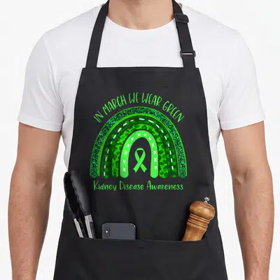 March kidney disease awareness apron - Green rainbow with awareness ribbon and text IN MARCH WE WEAR GREEN Kidney — shown on a black cotton canvas apron against a white background