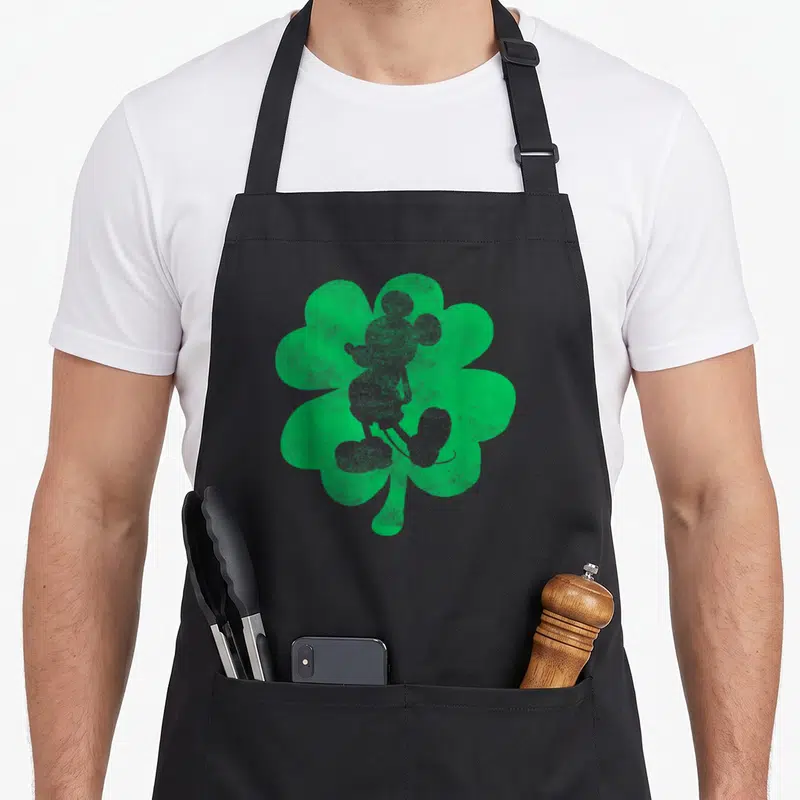 Shamrock mice looks apron - Grey Mickey Mouse silhouette inside a green distressed shamrock — shown on a black cotton canvas apron against a white background