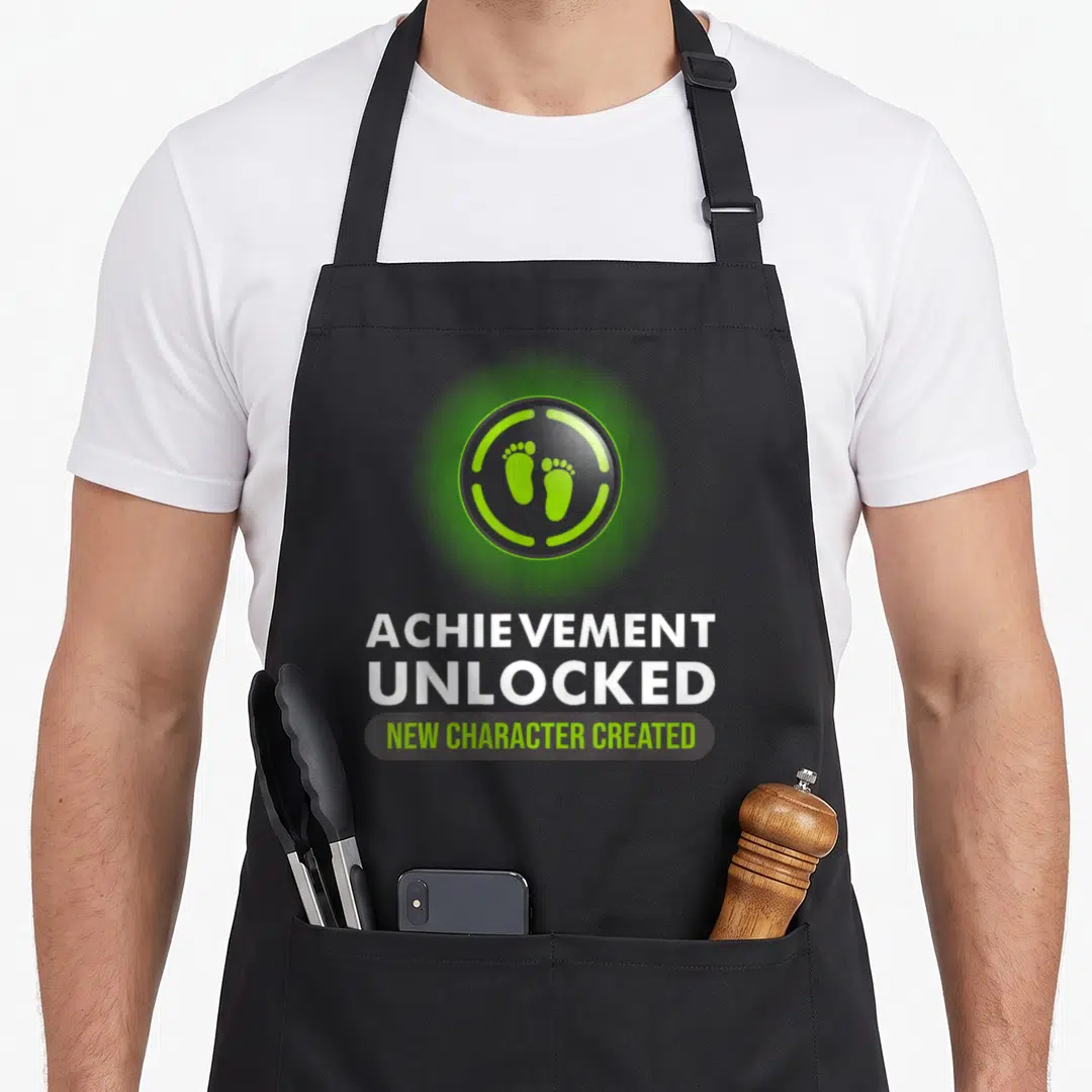 Pregnant character aprons - Green circular badge with two footprints above Achievement Unlocked New Character Created — shown on a black cotton canvas apron against a white background