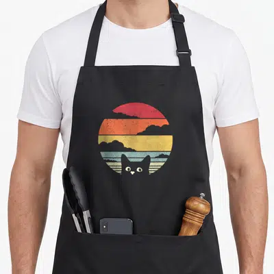Retro cat apron - Cat head peeking from red orange yellow blue striped sun with grey clouds — shown on a black cotton canvas apron against a white background