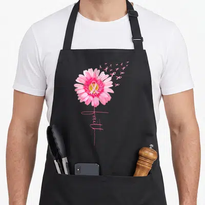 Daisy ribbon apron - Pink daisy with ribbon center and floating ribbons, faith script below — shown on a black cotton canvas apron against a white background