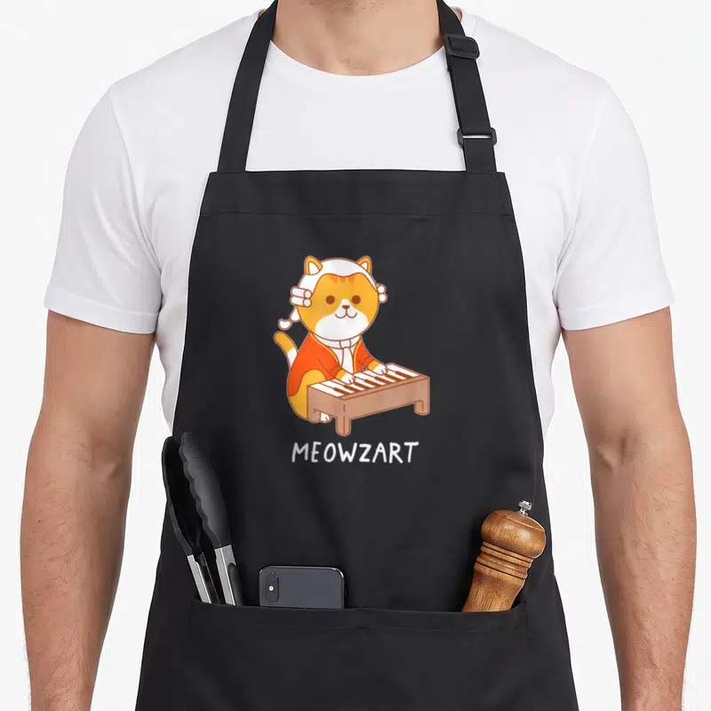Meowzart piano apron - Orange and white cartoon cat standing behind a keyboard with MEOWZART text below — shown on a black cotton canvas apron against a white background