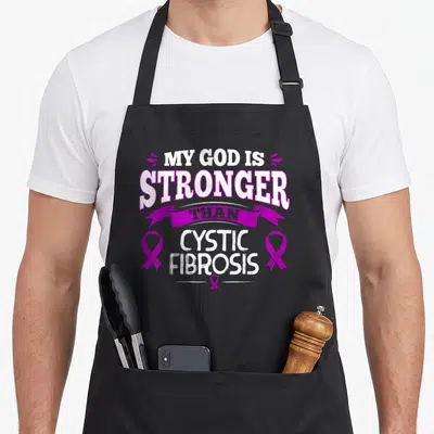 Awareness cystic ribbon apron - Purple and white My God Is Stronger Than Cystic Fibrosis text with ribbons — shown on a black cotton canvas apron against a white background