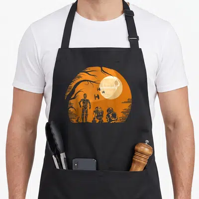 Droid halloween apron - Three droids and Death Star inside an orange circle with tree branches — shown on a black cotton canvas apron against a white background