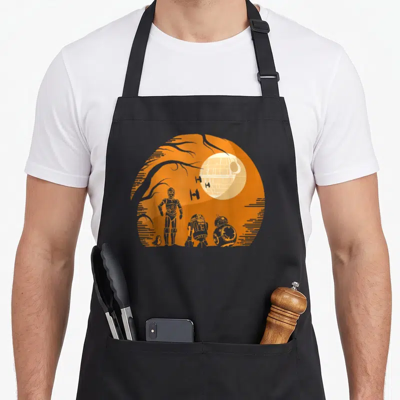 Droid halloween apron - Three droids and Death Star inside an orange circle with tree branches — shown on a black cotton canvas apron against a white background