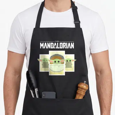 Panel cartoon apron - The Mandalorian text with Grogu in pod and two side figures in green and beige — shown on a black cotton canvas apron against a white background