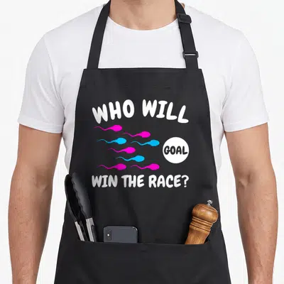 Win gender reveal apron - Cartoon pink and blue sperm swimming toward a white goal circle with text — shown on a black cotton canvas apron against a white background