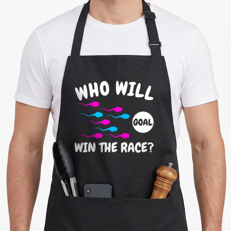 Win gender reveal apron - Cartoon pink and blue sperm swimming toward a white goal circle with text — shown on a black cotton canvas apron against a white background