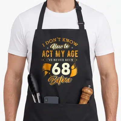 68th act birthday apron - Orange and white text reading I DON'T KNOW How to ACT MY AGE 68 Before with ribbon ends — shown on a black cotton canvas apron against a white background