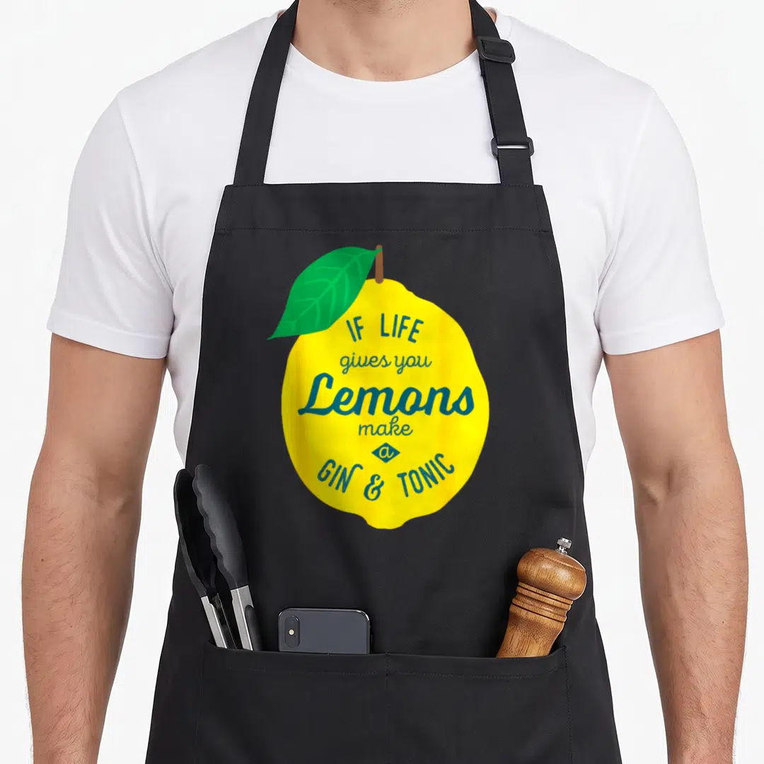 Gin lemon apron - Yellow lemon illustration with green leaf and blue text reading if life gives you lemons make a gin — shown on a black cotton canvas apron against a white background