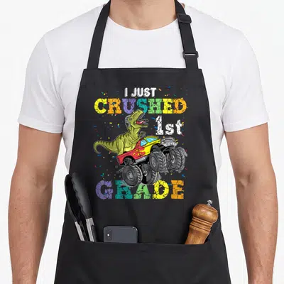Crushed 1st truck apron - Dinosaur in monster truck with I JUST CRUSHED 1st GRADE text and confetti — shown on a black cotton canvas apron against a white background