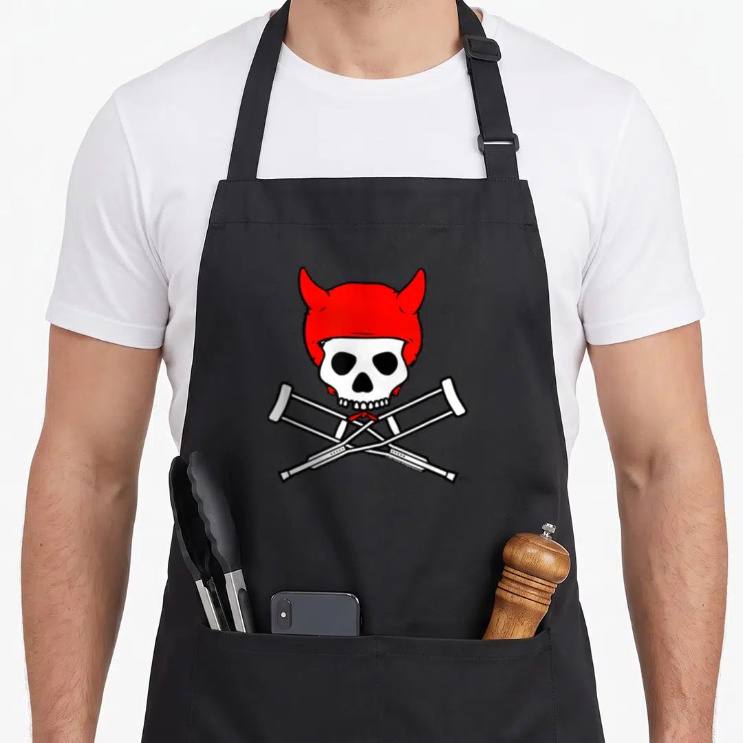 Devil skull apron - Cartoon skull with red devil horns above two crossed white crutches — shown on a black cotton canvas apron against a white background
