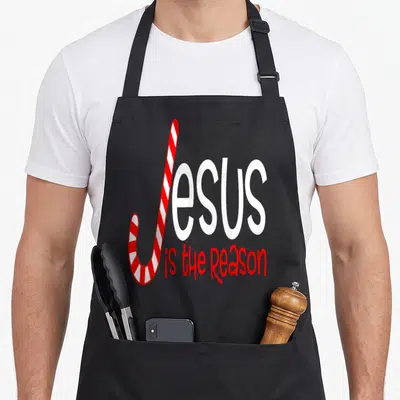 Jesus candy cane apron - Red and white candy cane forming J in Jesus is the Reason text — shown on a black cotton canvas apron against a white background